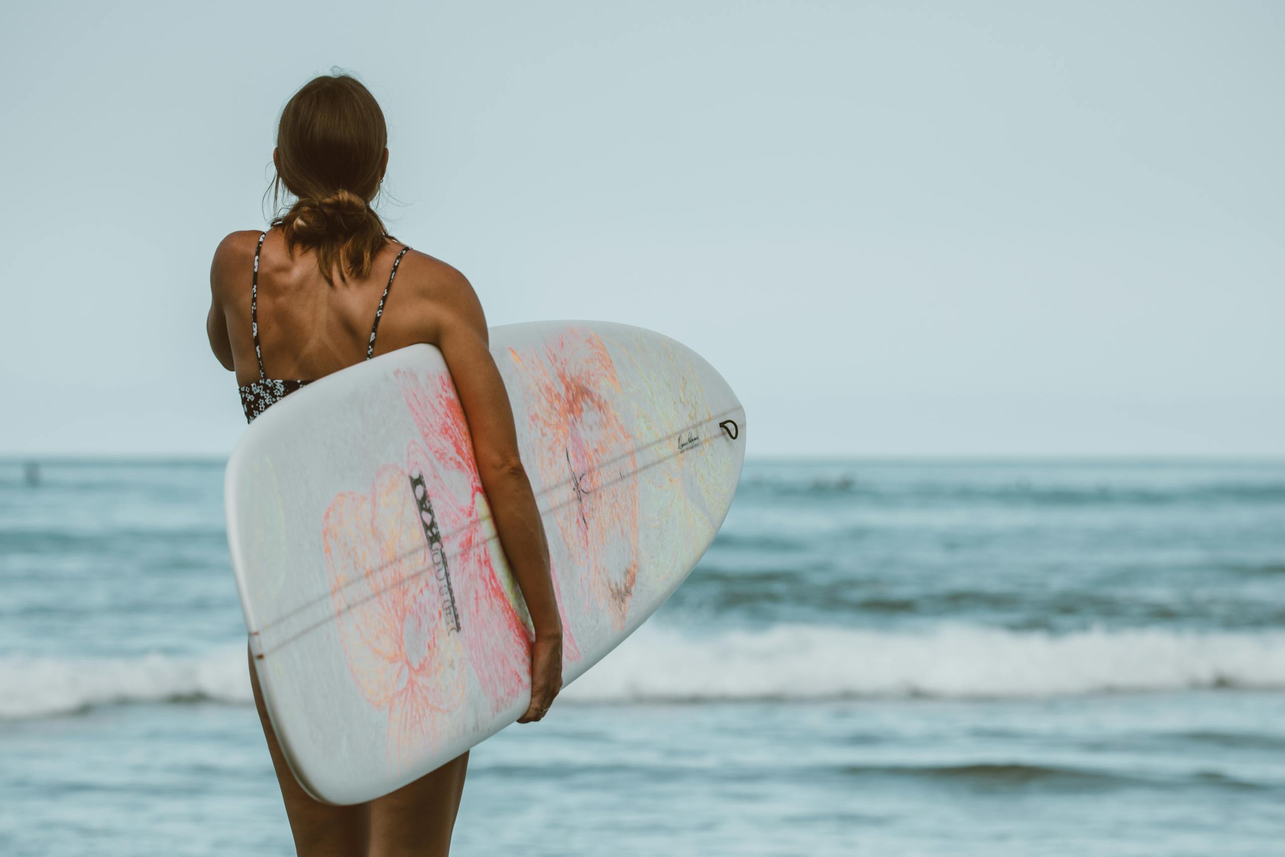 A woman holding a surfboard gazes at the ocean on a sunny Honolulu beach.