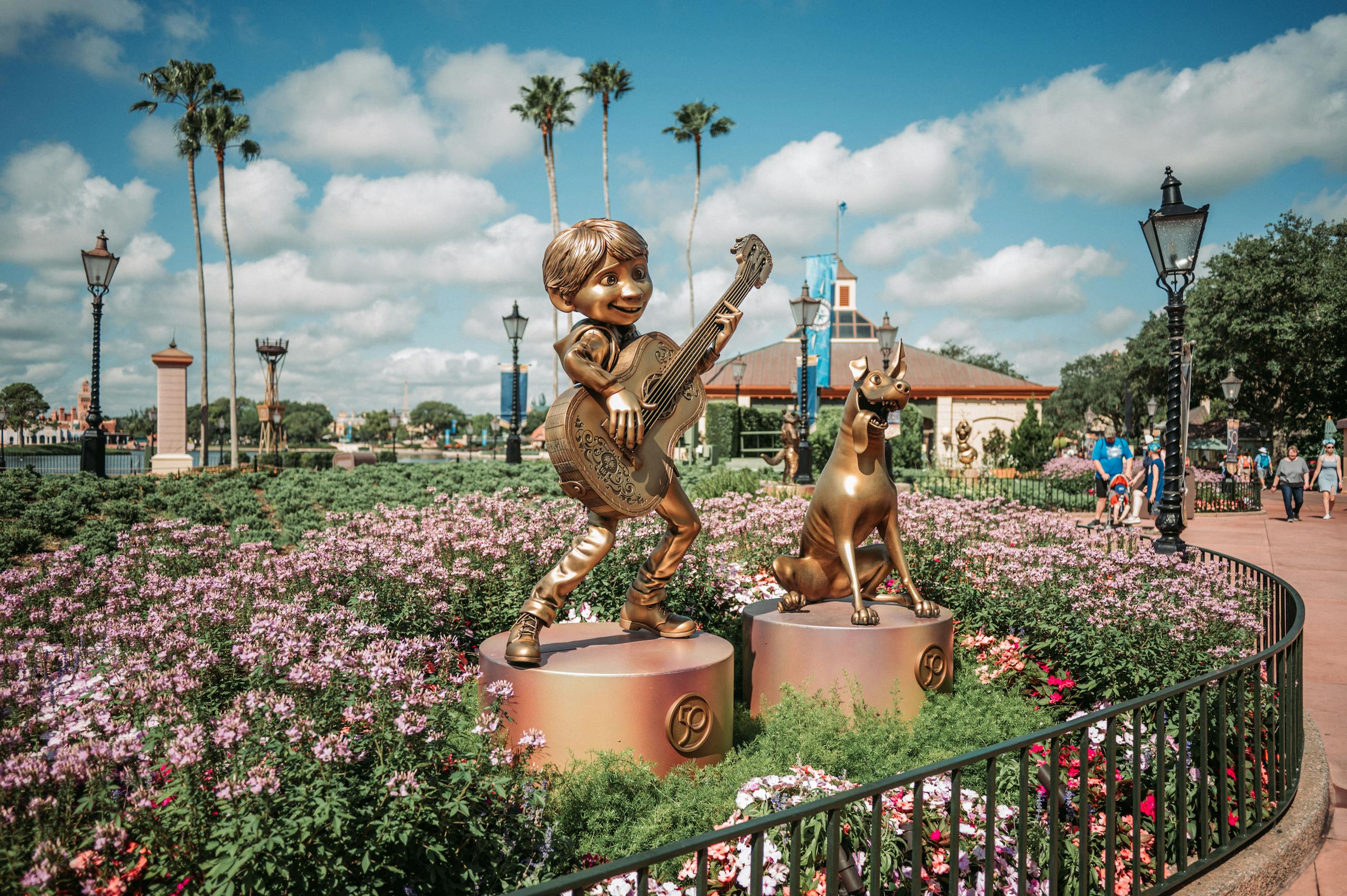 Golden character statues with guitar and dog surrounded by vibrant flowers in a theme park setting.