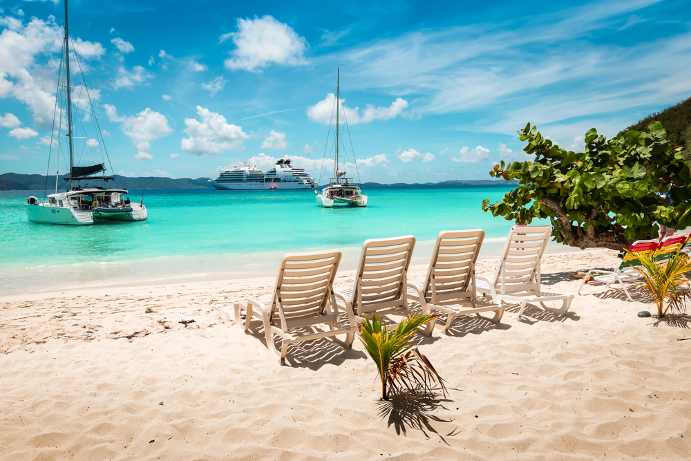 white-sand beach with 4 white chairs facing the ocean with large catamarans in the British Virgin Islands