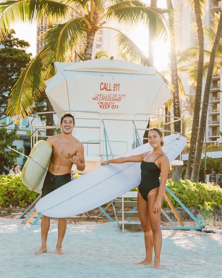 A cheerful couple poses with surfboards on a sunny beach, surrounded by palm trees and a lifeguard tower.