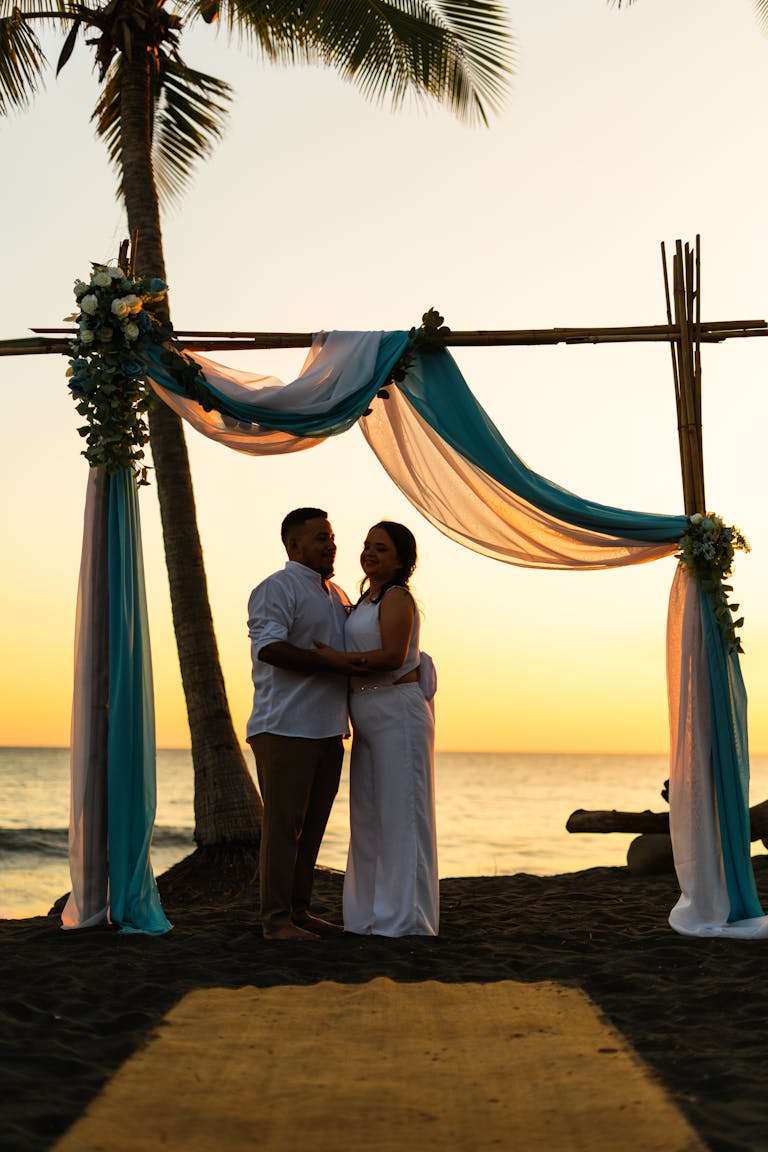 A romantic beach wedding ceremony with a couple embracing under a decorated arch at sunset.